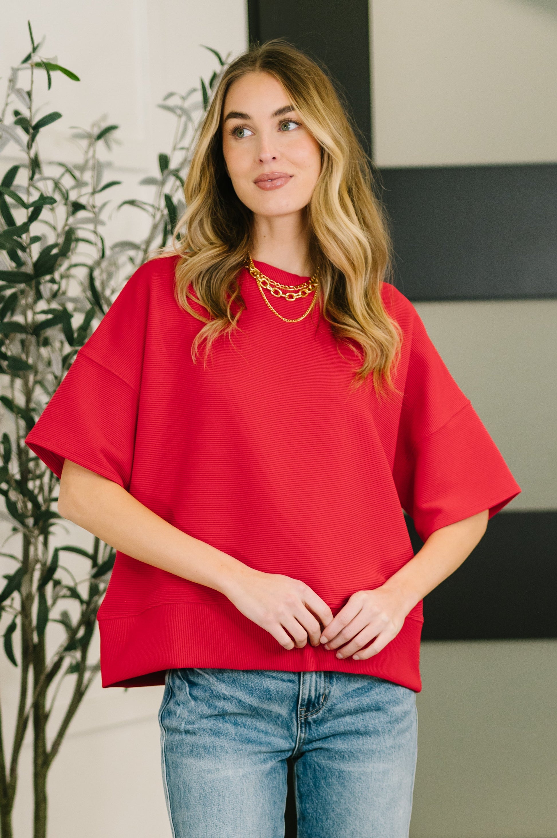 Woman wearing a red top and blue jeans standing indoors with a plant in the background