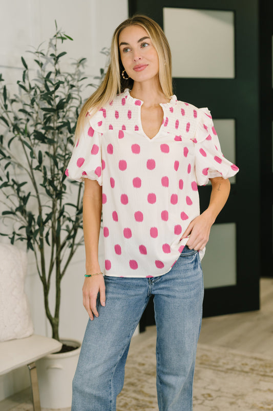 Woman wearing a white blouse with pink polka dots and blue jeans indoors.