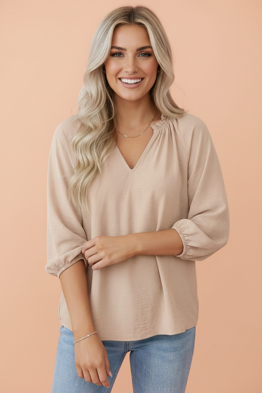 A smiling blonde woman wearing a light beige V-neck blouse with three-quarter sleeves and blue jeans, standing against a solid peach studio background.