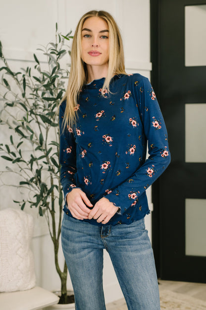 Woman wearing a blue floral sweater and jeans standing indoors with a plant in the background