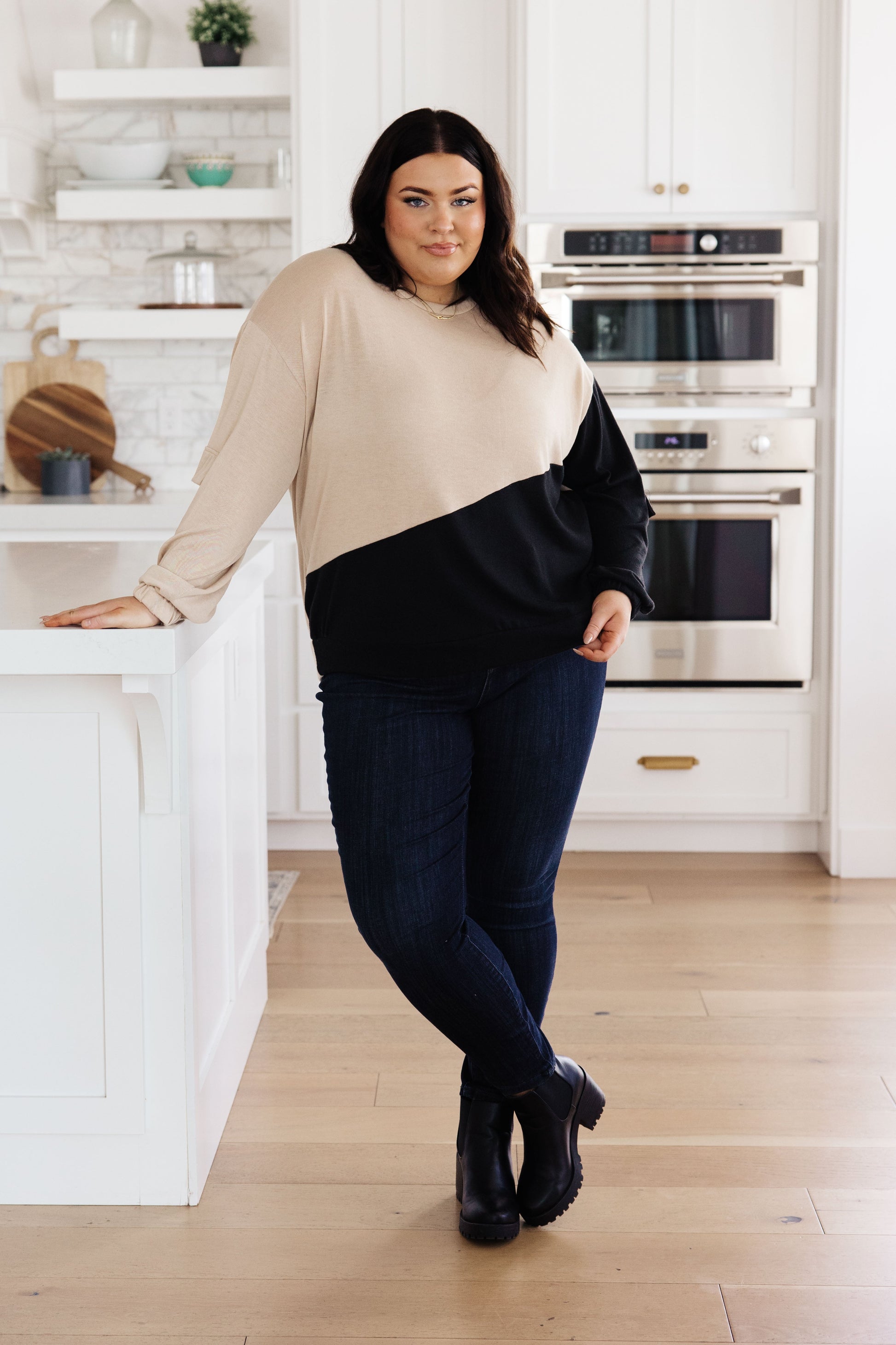 Woman standing in a kitchen wearing a beige and black sweater with blue jeans.