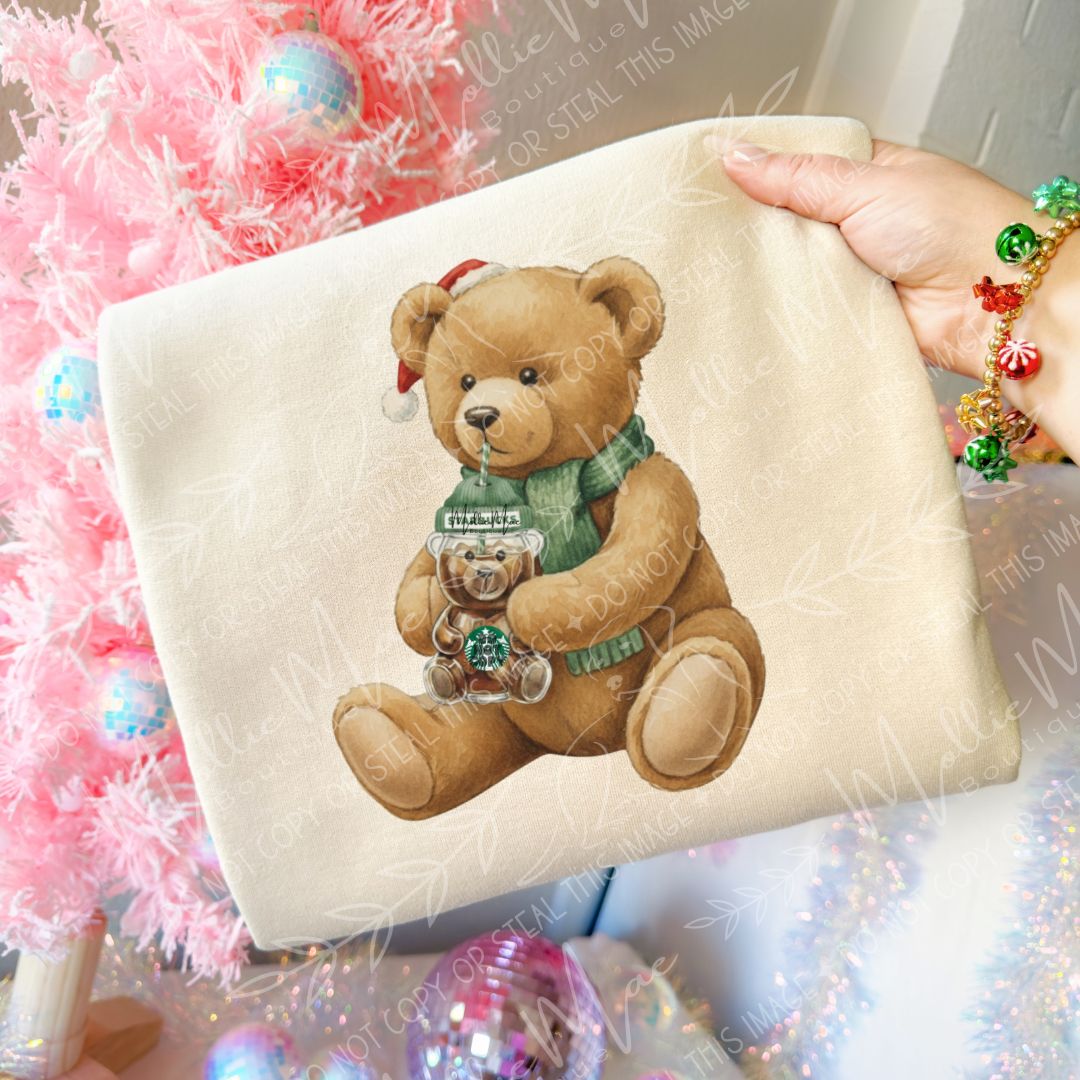 “Folded Gildan Sand sweatshirt with Starbs Bear graphic design showing a teddy bear sipping a festive drink. Pink Christmas tree and ornaments in background.”
