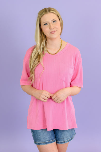 Woman wearing a pink top and denim shorts indoors with a plant in the background