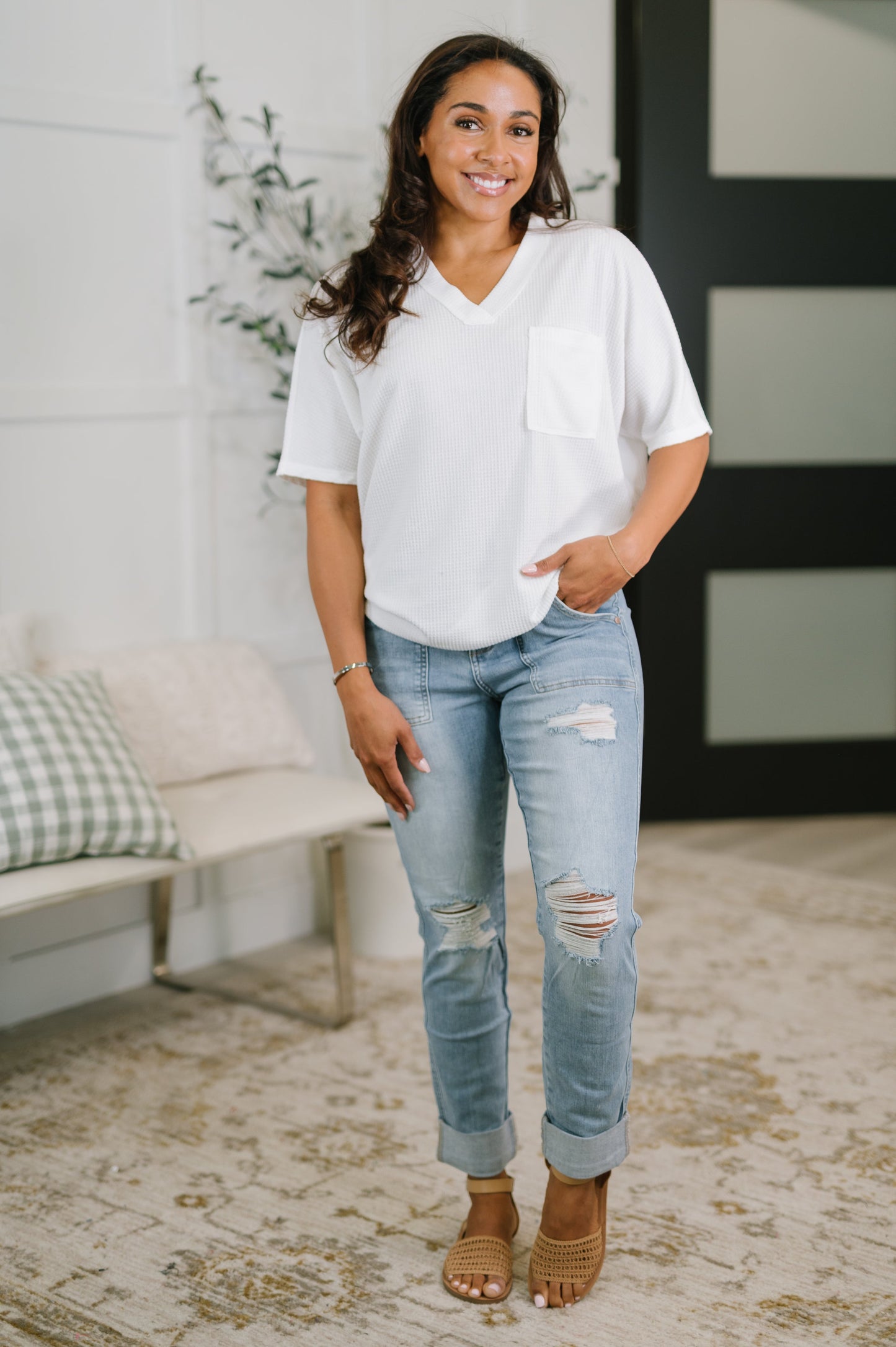 Woman wearing a white blouse and light blue jeans standing in a room with a couch and plant.