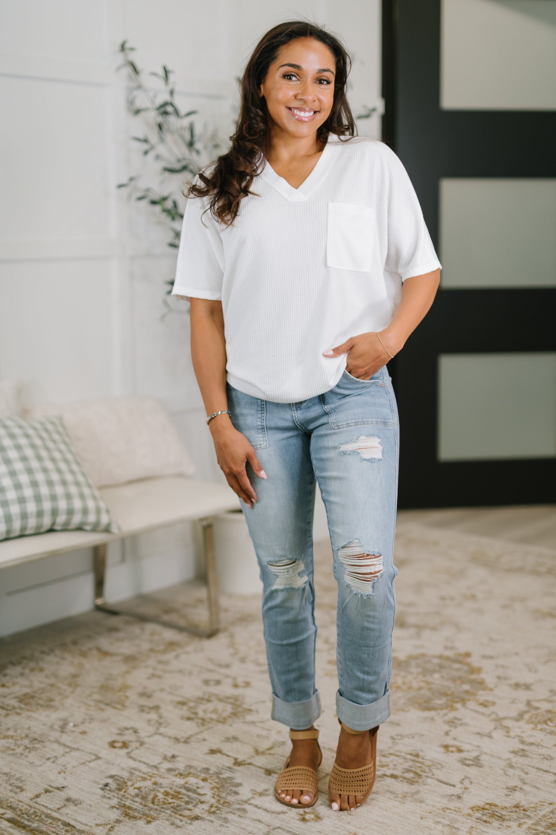 Woman wearing a white blouse and light blue jeans standing in a room with a couch and plant.