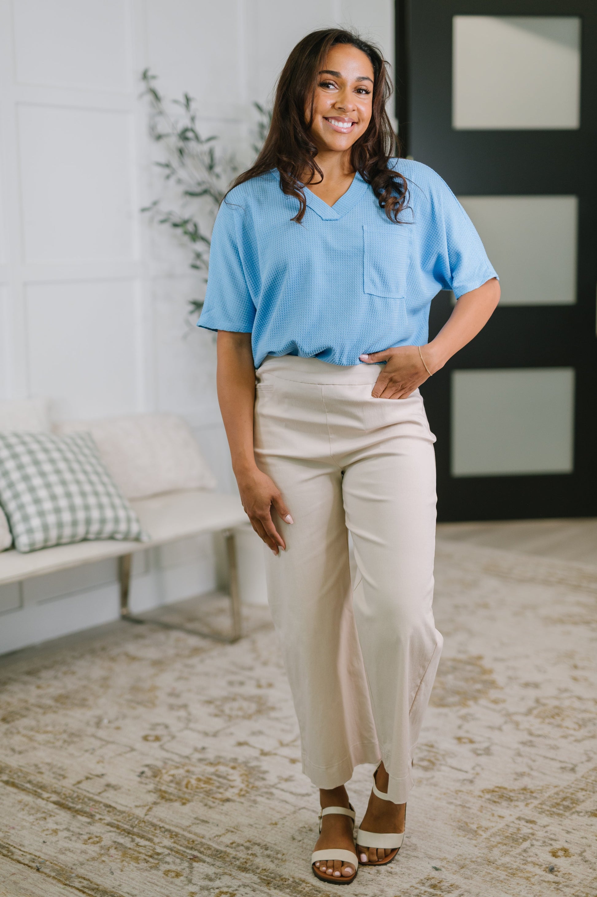 Woman wearing a blue top and beige pants standing in a room with a white wall and a plant.