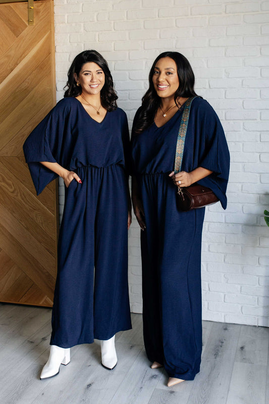 Two women wearing navy blue dresses standing in front of a wooden door and white brick wall.