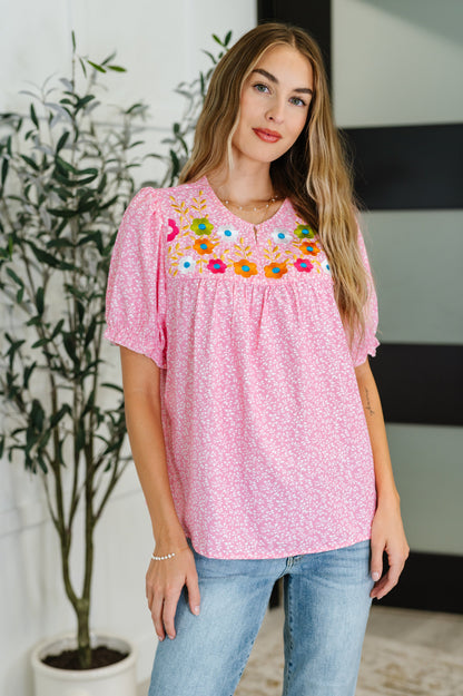 Woman wearing a pink blouse with floral embroidery in an indoor setting.