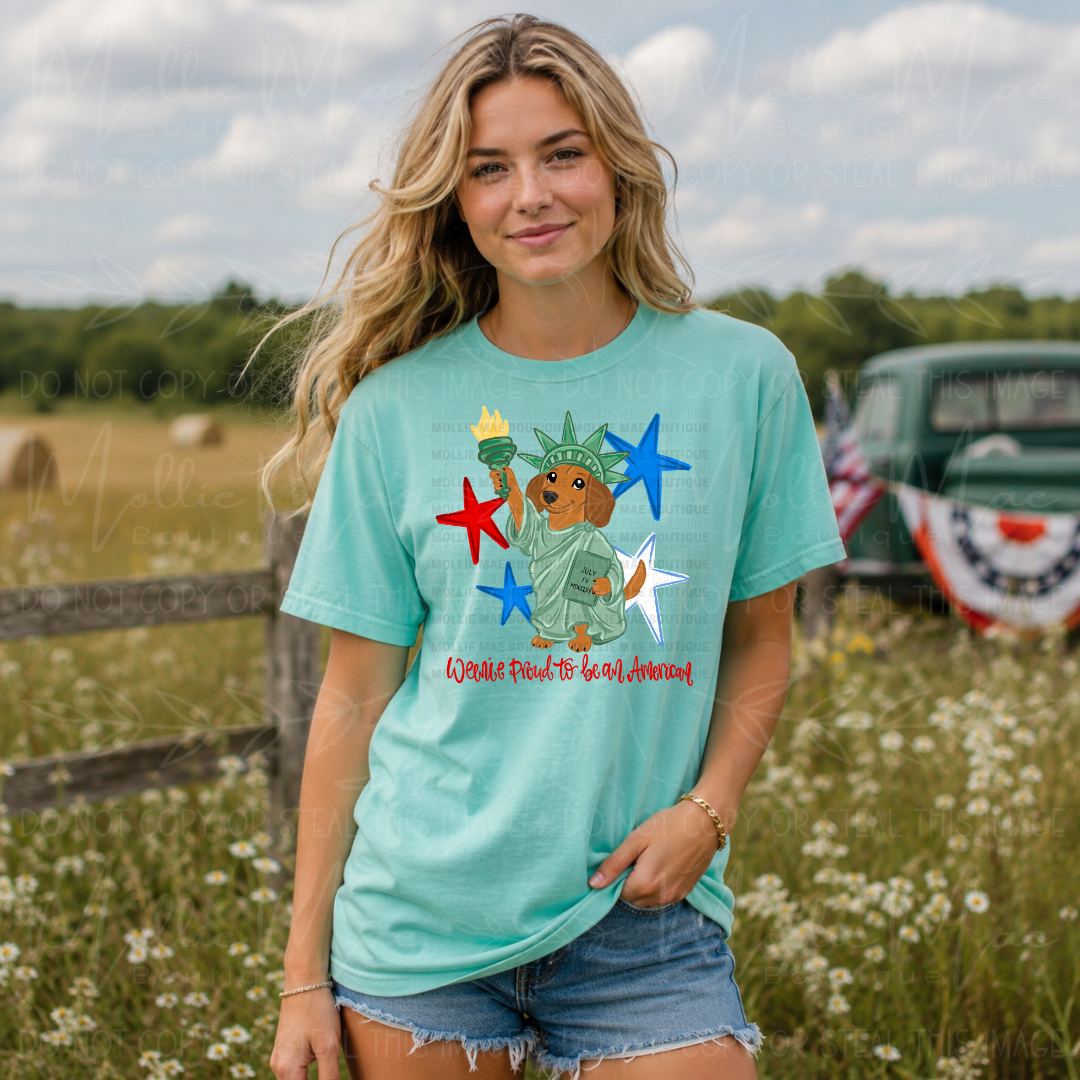 Comfort Colors Chalky Mint graphic tee featuring a patriotic dachshund dressed as the Statue of Liberty with red, white, and blue stars and optional Weenie Proud to Be American wording.