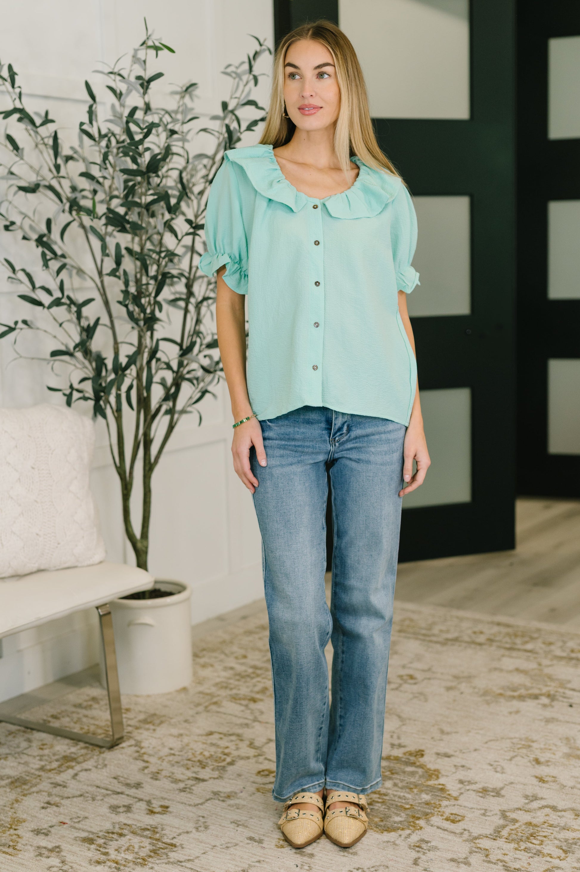 Woman wearing a light blue blouse and jeans standing in a room with a plant and sofa.