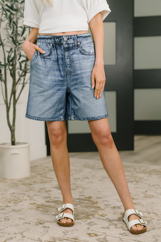 Person wearing blue denim shorts standing indoors with a plant and wall in the background