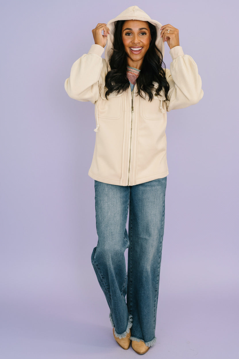 Woman wearing a cream jacket and blue jeans standing indoors next to a plant.